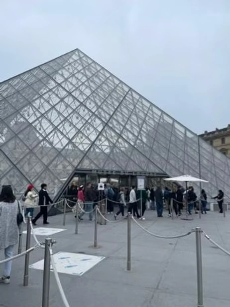 A large glass pyramid entrance with visitors lining up in an overcast plaza.